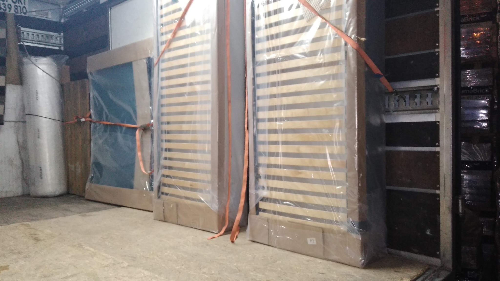 Łóżka i inne meble zabezpieczone pasami w przestrzeni ładunkowej. Elementy owinięte folią ochronną przed transportem. Widok na załadunek wewnątrz pojazdu.