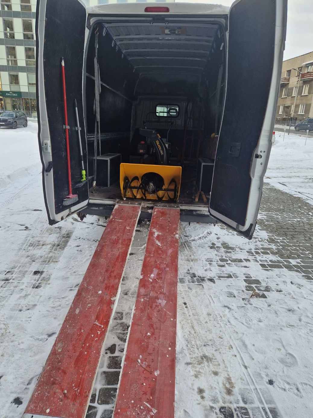 Otwarty bus z rampami i odśnieżarką w środku, gotowy do transportu zimą. Na drzwiach zamontowane narzędzia.
