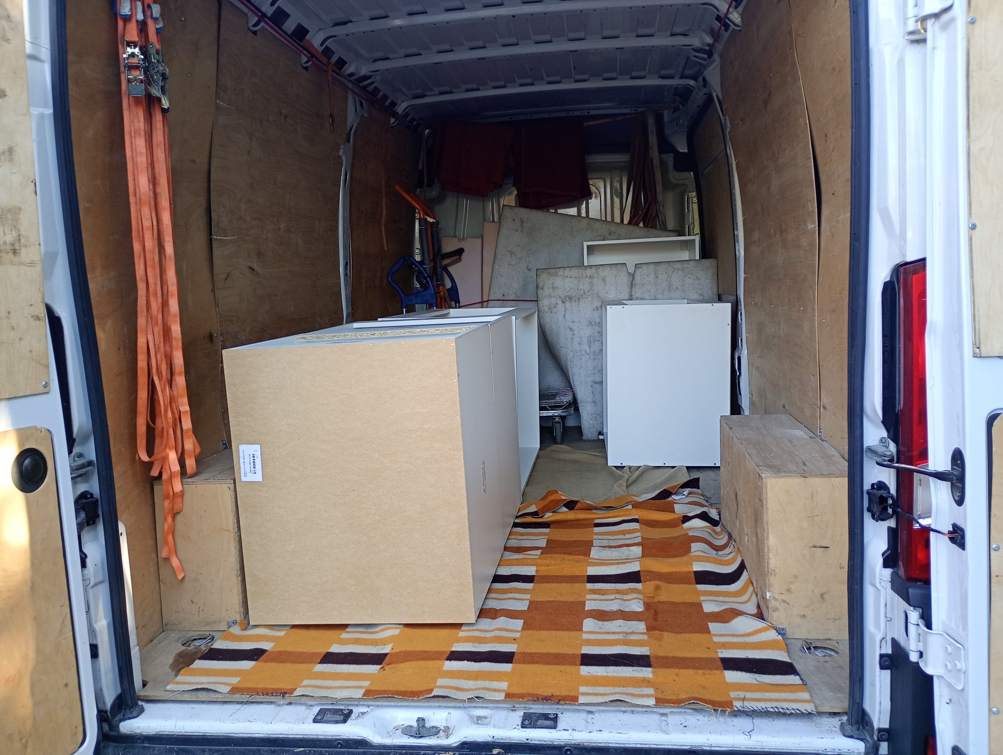 Wnętrze białego busa załadowane meblami i zabezpieczone pasami transportowymi. Widoczny dywan na podłodze, kartony i szafki gotowe do przewozu.