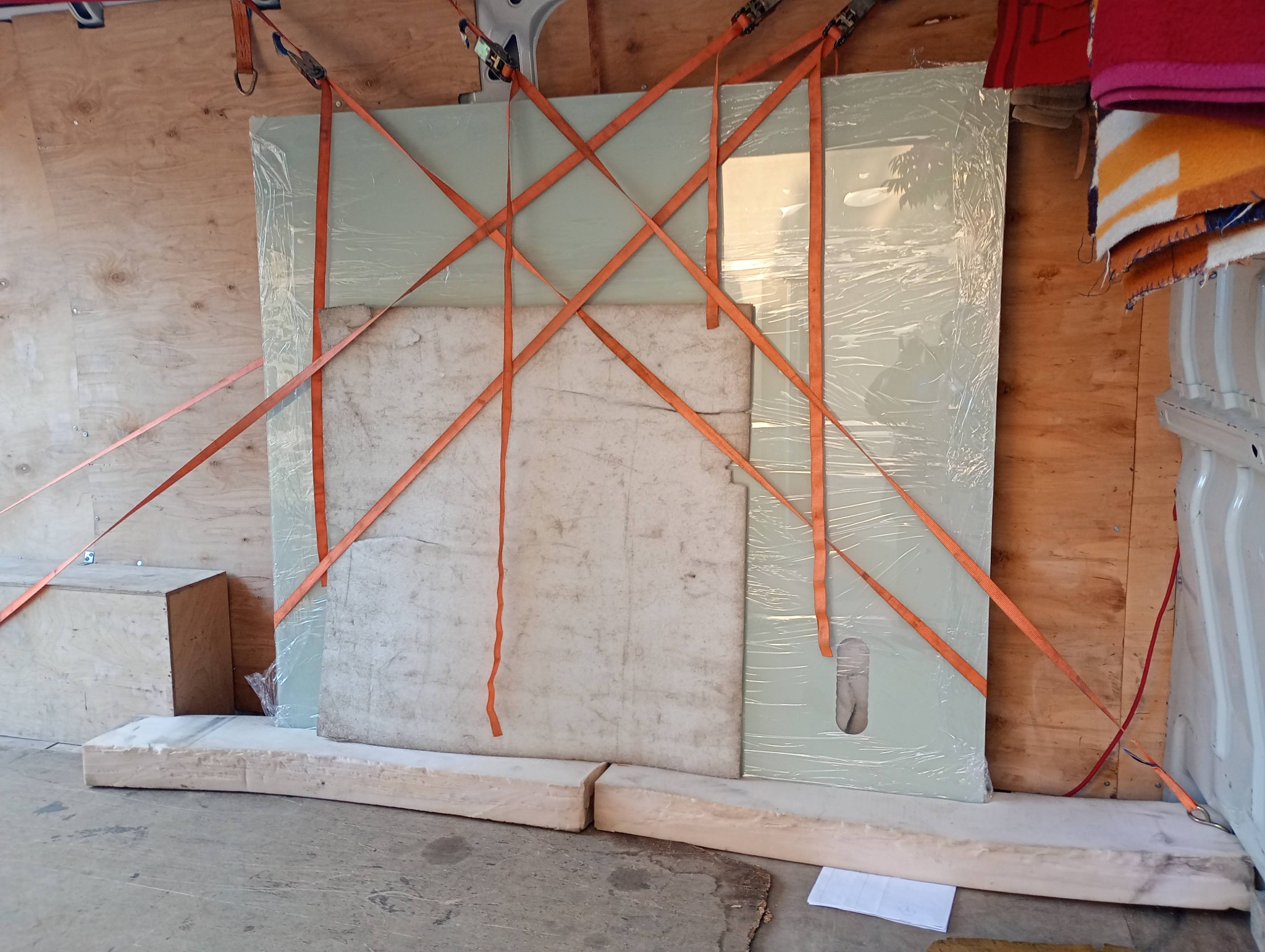 Płyty zabezpieczone folią i pasami transportowymi wewnątrz pojazdu. Drewniana skrzynia i koce widoczne w tle. Bezpieczny transport ładunku.