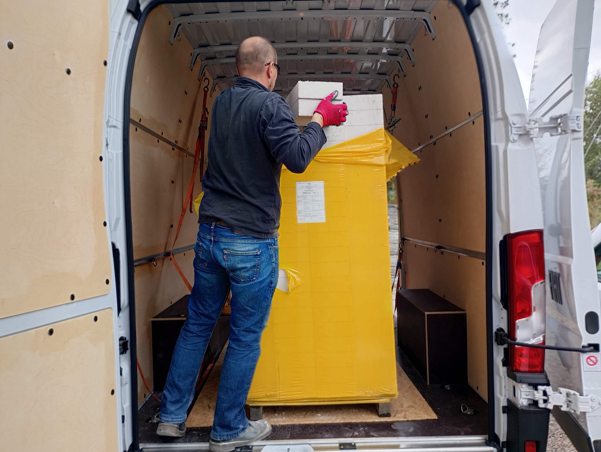 Pracownik w czerwonych rękawicach umieszcza białe płyty na żółtej, zabezpieczonej paczce wewnątrz furgonetki z drewnianą zabudową i otwartymi drzwiami.