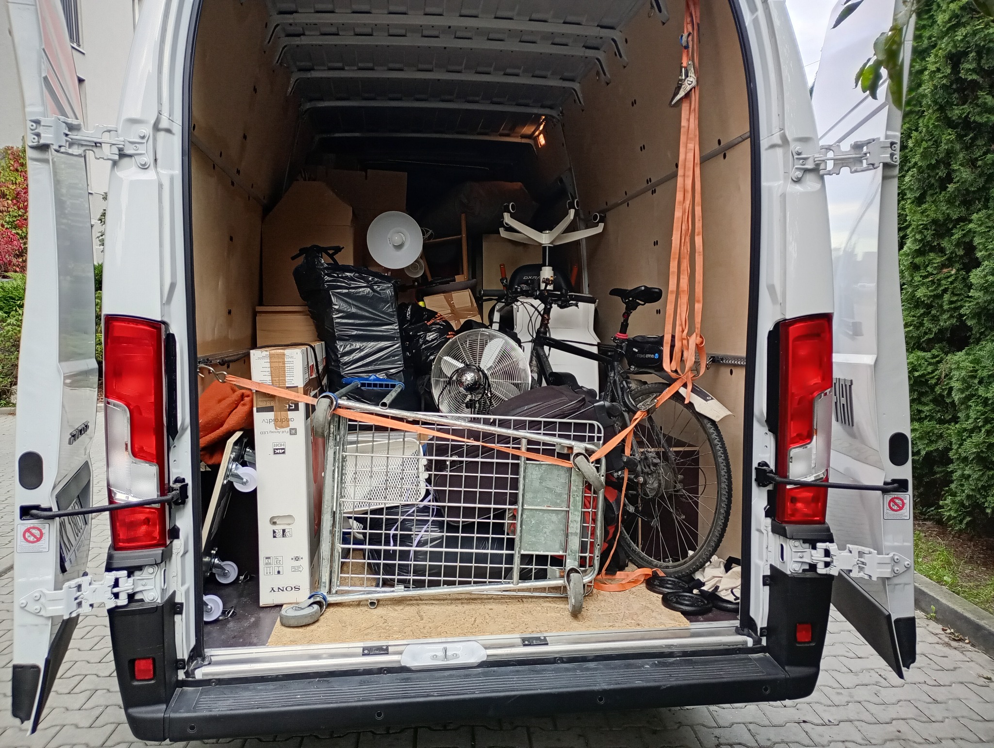Wnętrze białego busa załadowanego różnymi przedmiotami, zabezpieczonymi pasami transportowymi, gotowego do transportu. Widoczny rower, krzesło biurowe, wentylator i kartony.