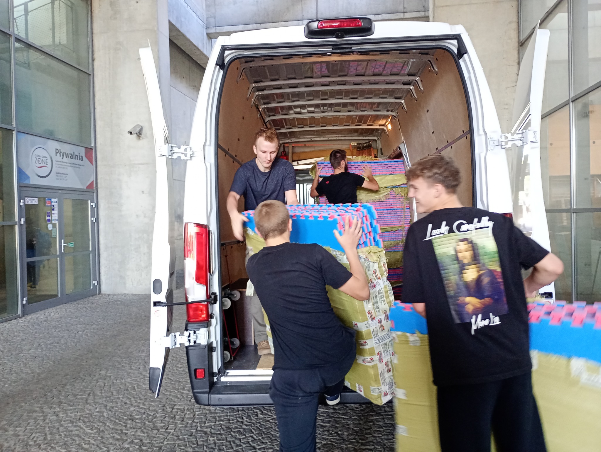 Ekipa ładuje do busa zapakowane maty piankowe na tle wejścia do pływalni. Widoczne otwarte drzwi busa i trzech mężczyzn w trakcie załadunku.