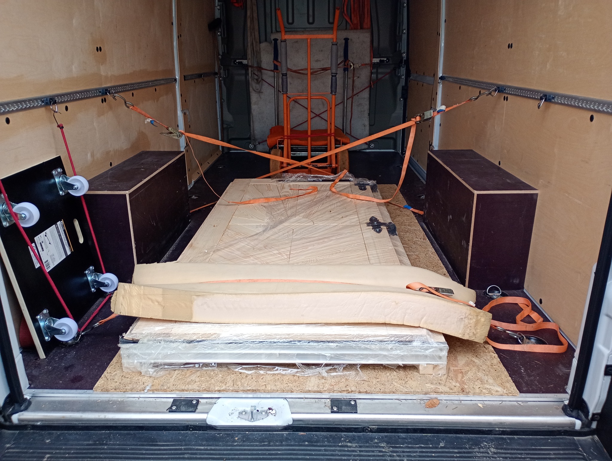 Wnętrze busa z zabezpieczonym ładunkiem: drzwi, wózek transportowy i platforma na kółkach. Pasy mocujące zapewniają bezpieczeństwo transportu.