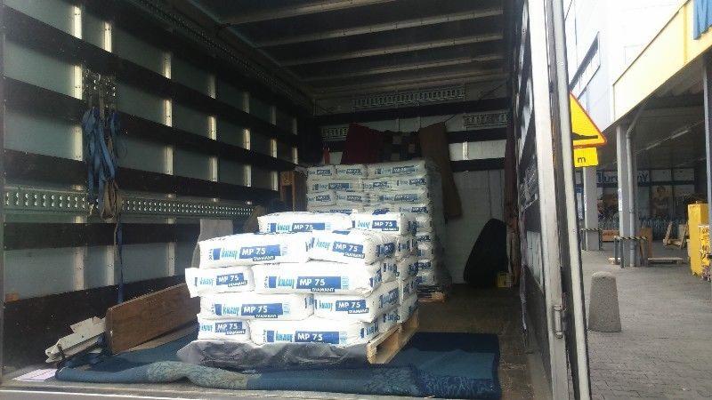 Wnętrze busa transportowego załadowane paletami z workami zaprawy murarskiej Knauf MP 75, widoczne zabezpieczenia ładunku i fragment chodnika na zewnątrz.