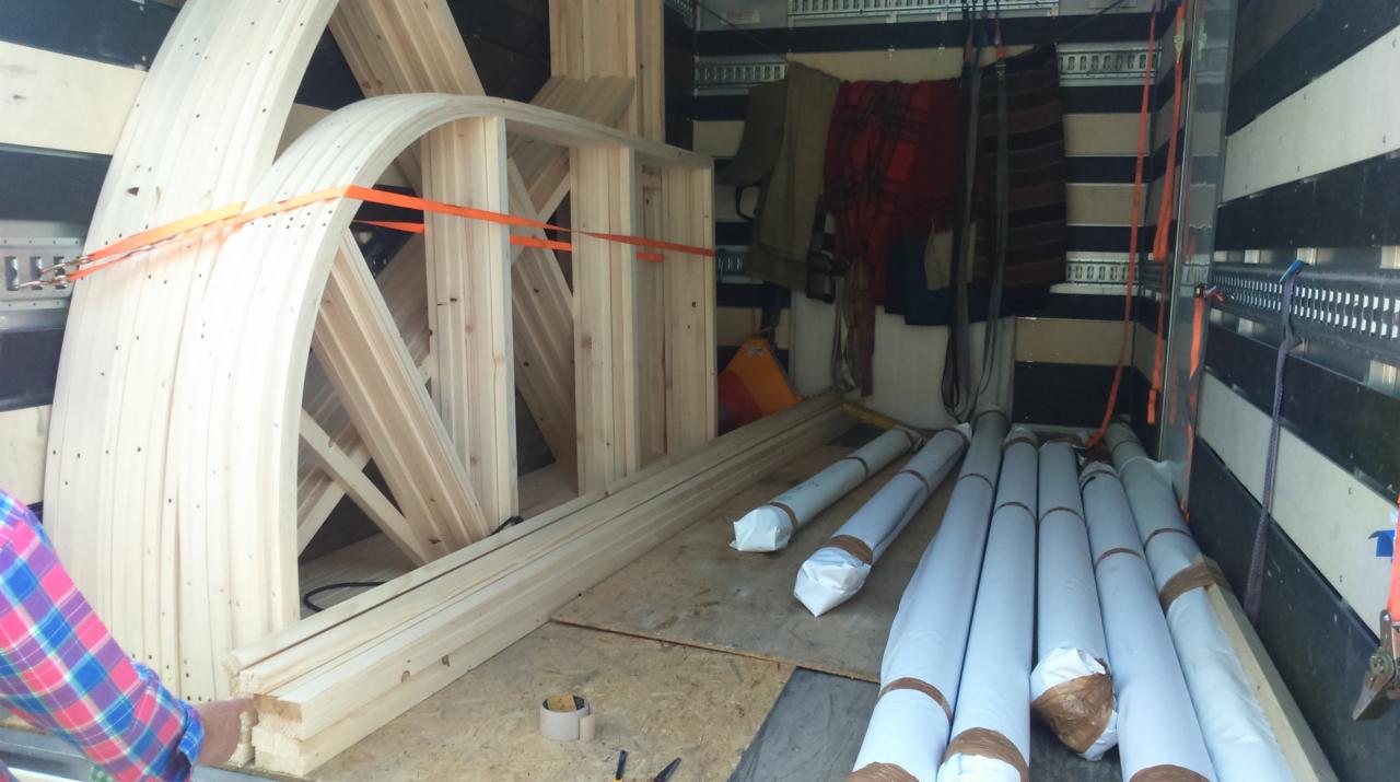 Wnętrze furgonetki załadowane elementami drewnianymi w kształcie łuków, zabezpieczonymi pomarańczowymi pasami, oraz zrolowanymi materiałami budowlanymi w białym opakowaniu.