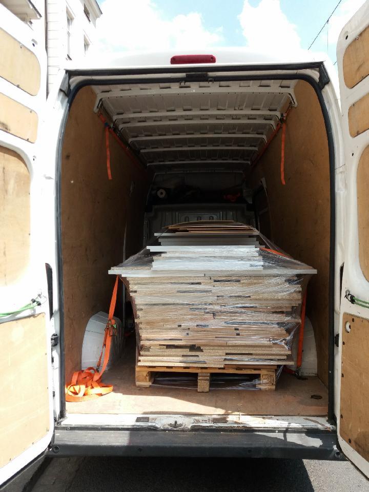 Wnętrze białego busa z otwartymi drzwiami, załadowane paletą z płytami zabezpieczonymi folią stretch i pasami mocującymi.