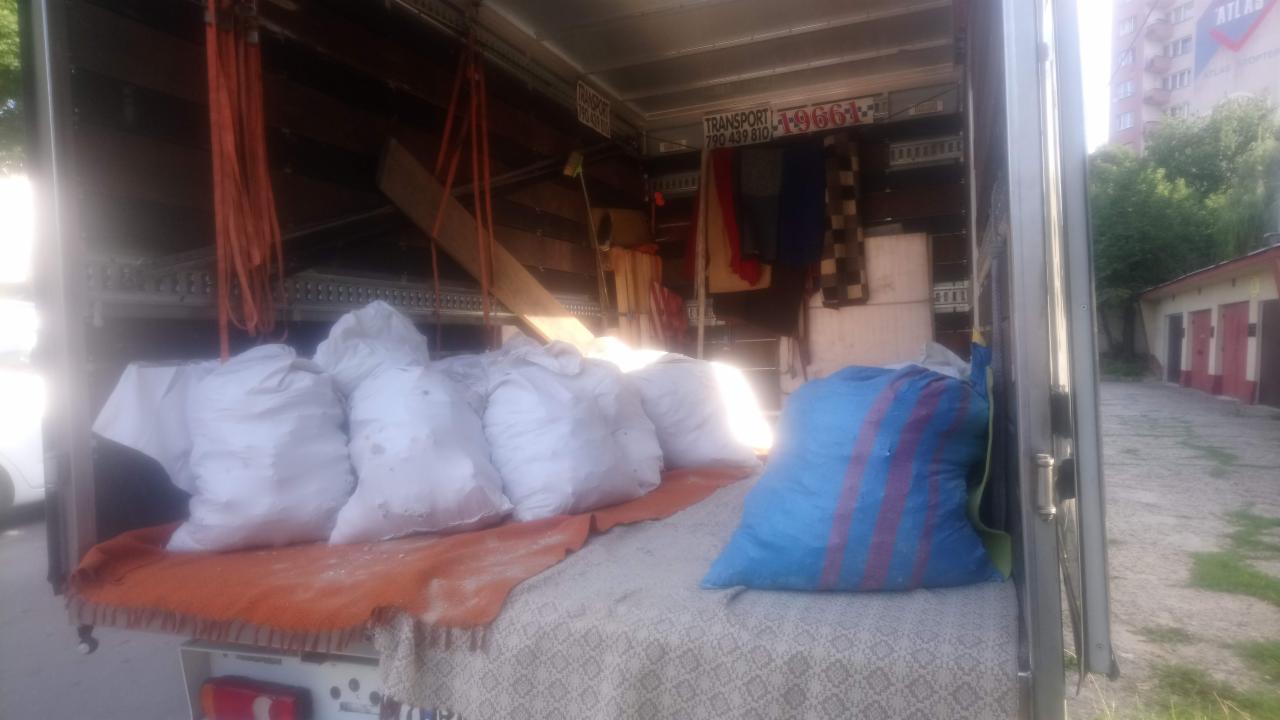 Wnętrze częściowo załadowanego busa transportowego: białe worki i niebieska torba w paski na pledzie i pomarańczowym kocu, widoczne pasy mocujące i tabliczka z numerem telefonu.