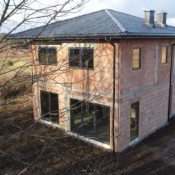 Surowy, dwukondygnacyjny dom z cegły ceramicznej w trakcie budowy, z zamontowanymi oknami i dachem z dachówki, widziany z perspektywy lotu ptaka w jesiennym krajobrazie.