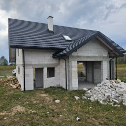 JNS SPÓŁKA Z OGRANICZONĄ ODPOWIEDZIALNOŚCIĄ - Surowy dom z białego betonu komórkowego z czarnym dachem, widoczne otwory okienne i drzwiowe, stosy materiałów budowlanych na zewnątrz, w tle zieleń.