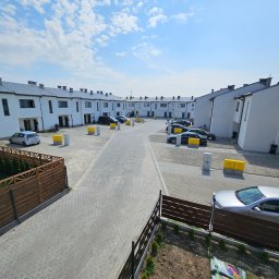 JNS SPÓŁKA Z OGRANICZONĄ ODPOWIEDZIALNOŚCIĄ - Osiedle domów jednorodzinnych w zabudowie szeregowej, z szarymi dachami i białymi elewacjami, widziane z góry. Na pierwszym planie drewniany płot i fragment trawnika.