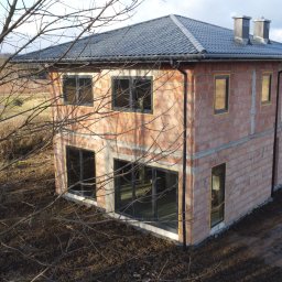 JNS SPÓŁKA Z OGRANICZONĄ ODPOWIEDZIALNOŚCIĄ - Surowy, dwukondygnacyjny dom z czerwonej cegły w trakcie budowy, z czarnymi ramami okiennymi, na tle jesiennego krajobrazu. Widoczne drzewa bez liści.