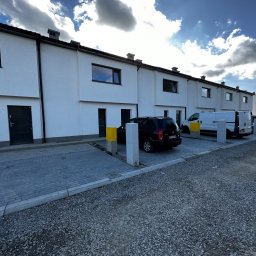 JNS SPÓŁKA Z OGRANICZONĄ ODPOWIEDZIALNOŚCIĄ - Rząd nowych, białych domów szeregowych z czarnymi oknami i drzwiami, zaparkowane samochody, kostka brukowa i gruz na podłożu, błękitne niebo z chmurami.