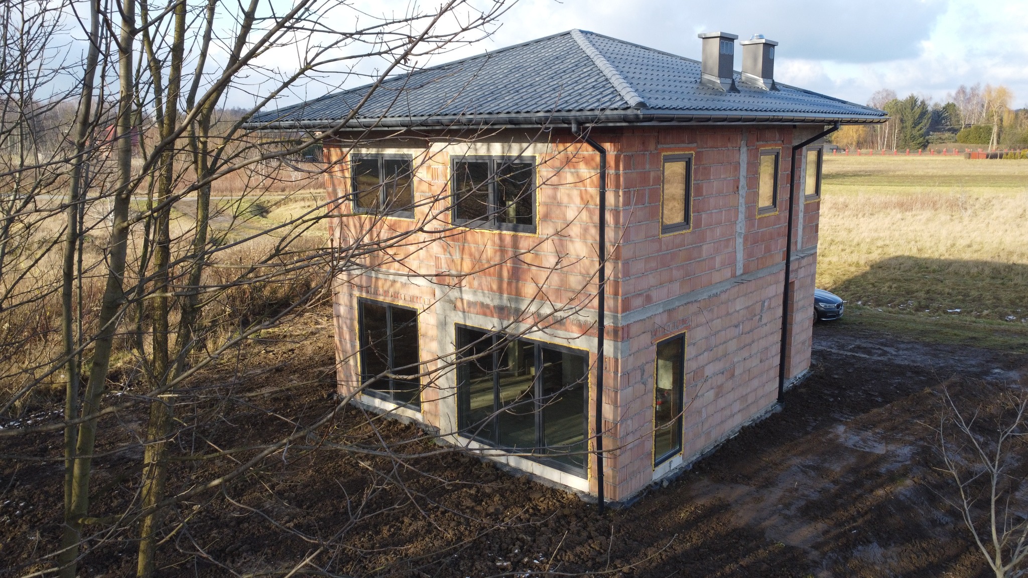 Surowy, dwukondygnacyjny dom z cegły ceramicznej w trakcie budowy, z zamontowanymi oknami i dachem z dachówki, widziany z perspektywy lotu ptaka w jesiennym krajobrazie.