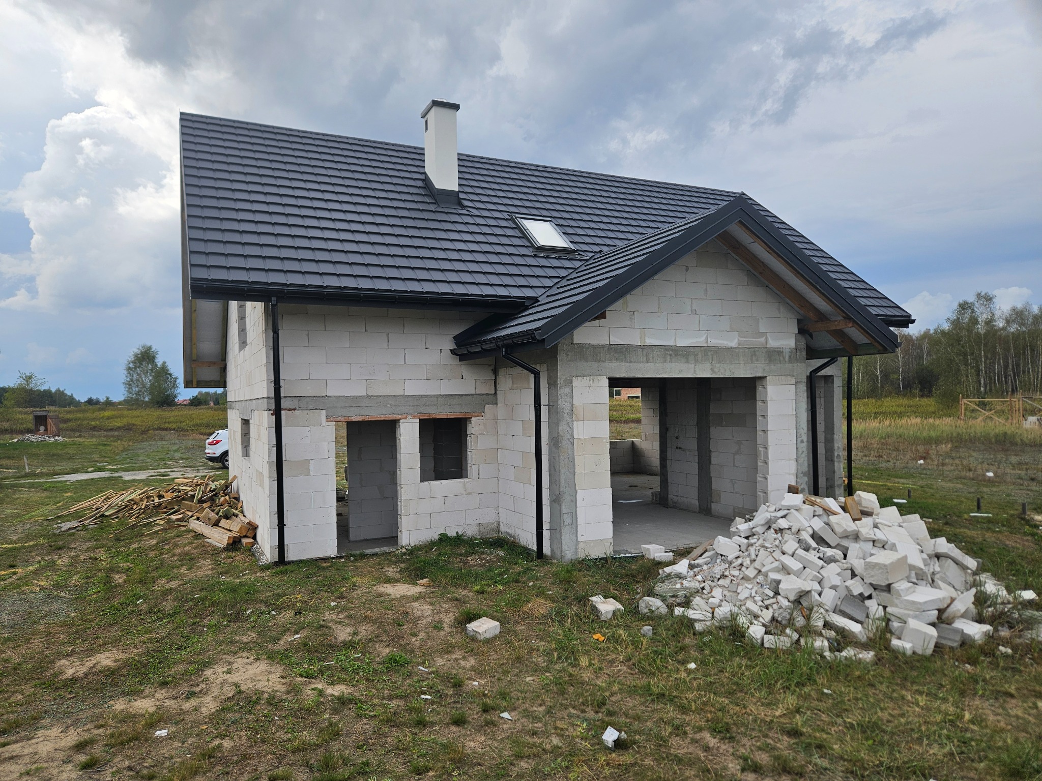 Surowy dom z białego betonu komórkowego z czarnym dachem, widoczne otwory okienne i drzwiowe, stosy materiałów budowlanych na zewnątrz, w tle zieleń.