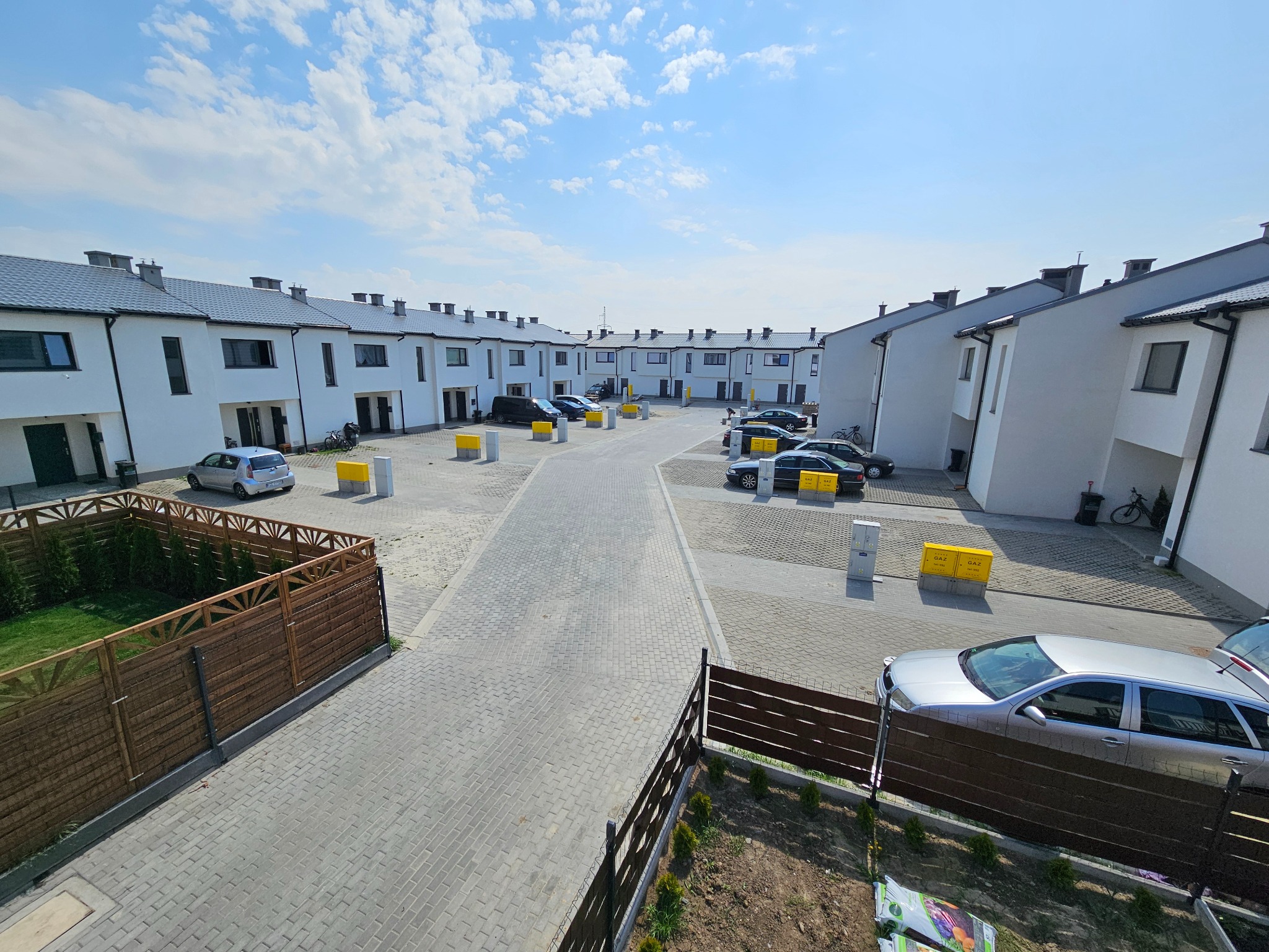 Osiedle domów jednorodzinnych w zabudowie szeregowej, z szarymi dachami i białymi elewacjami, widziane z góry. Na pierwszym planie drewniany płot i fragment trawnika.