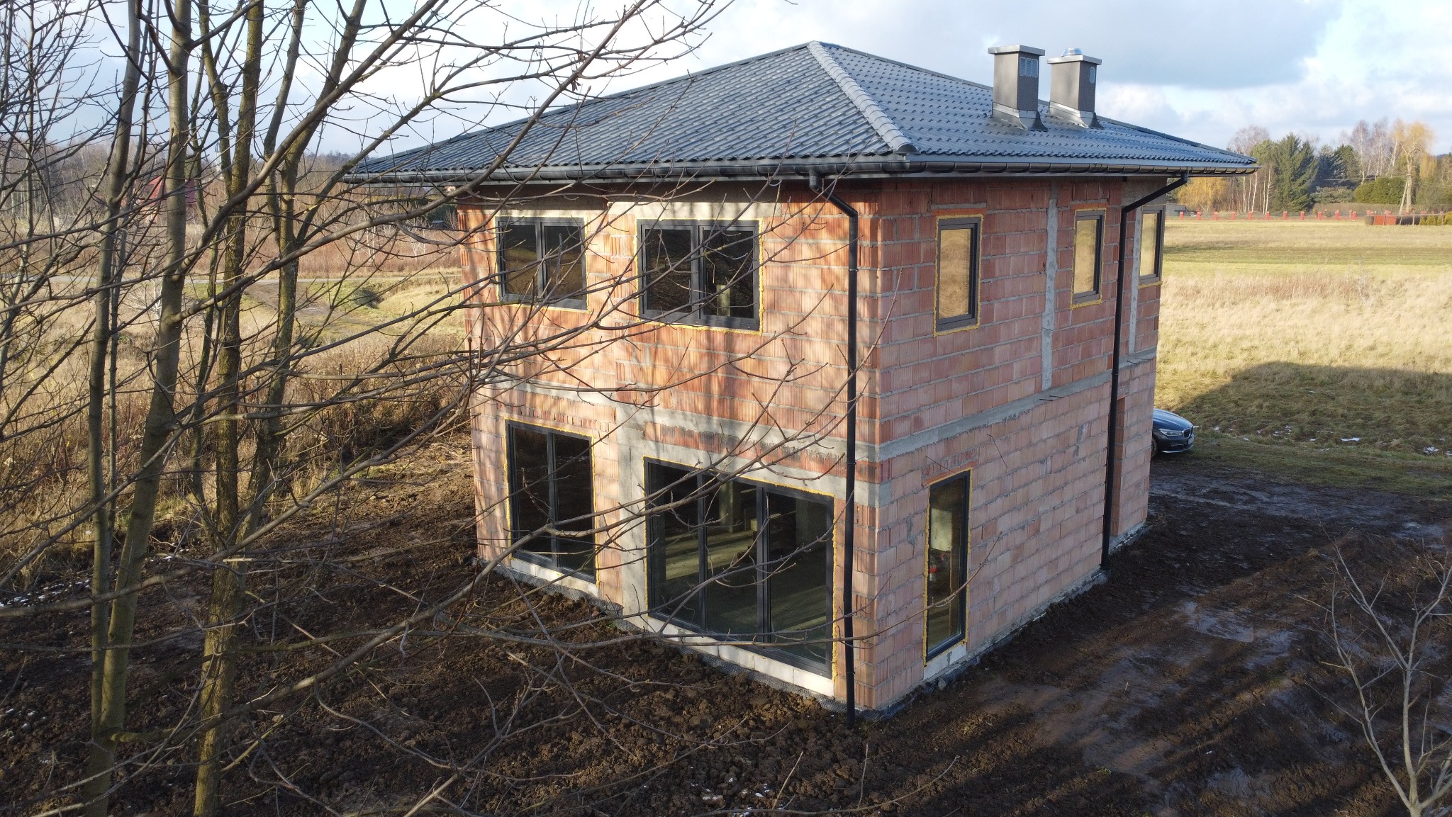 Surowy, dwukondygnacyjny dom z czerwonej cegły w trakcie budowy, z czarnymi ramami okiennymi, na tle jesiennego krajobrazu. Widoczne drzewa bez liści.