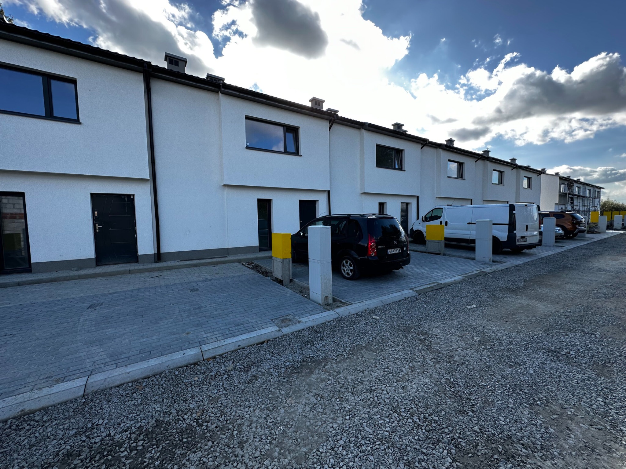 Rząd nowych, białych domów szeregowych z czarnymi oknami i drzwiami, zaparkowane samochody, kostka brukowa i gruz na podłożu, błękitne niebo z chmurami.