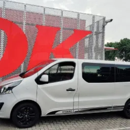 Biały bus marki Opel Vivaro z czarnymi felgami i paskami dekoracyjnymi, zaparkowany na tle nowoczesnego budynku z czerwonym napisem 'OK'.