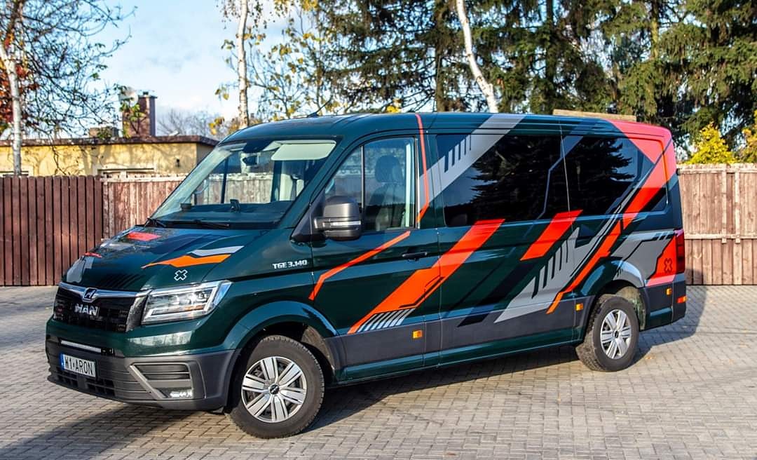 Zielony bus MAN TGE 3.140 z dynamicznym pomarańczowo-szarym wzorem na karoserii, zaparkowany na kostce brukowej przed budynkiem i drzewami.