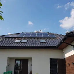 Nowoczesny dom jednorodzinny z ciemną dachówką i panelami fotowoltaicznymi na dachu, widok z dołu na błękitne niebo z chmurami.