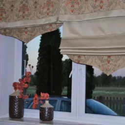 Beżowe zasłony rzymskie i dekoracyjne firany z motywem roślinnym w oknie z widokiem na ogród i samochód, uzupełnione dwoma brązowymi wazonami z pomarańczowymi kwiatami na parapecie.