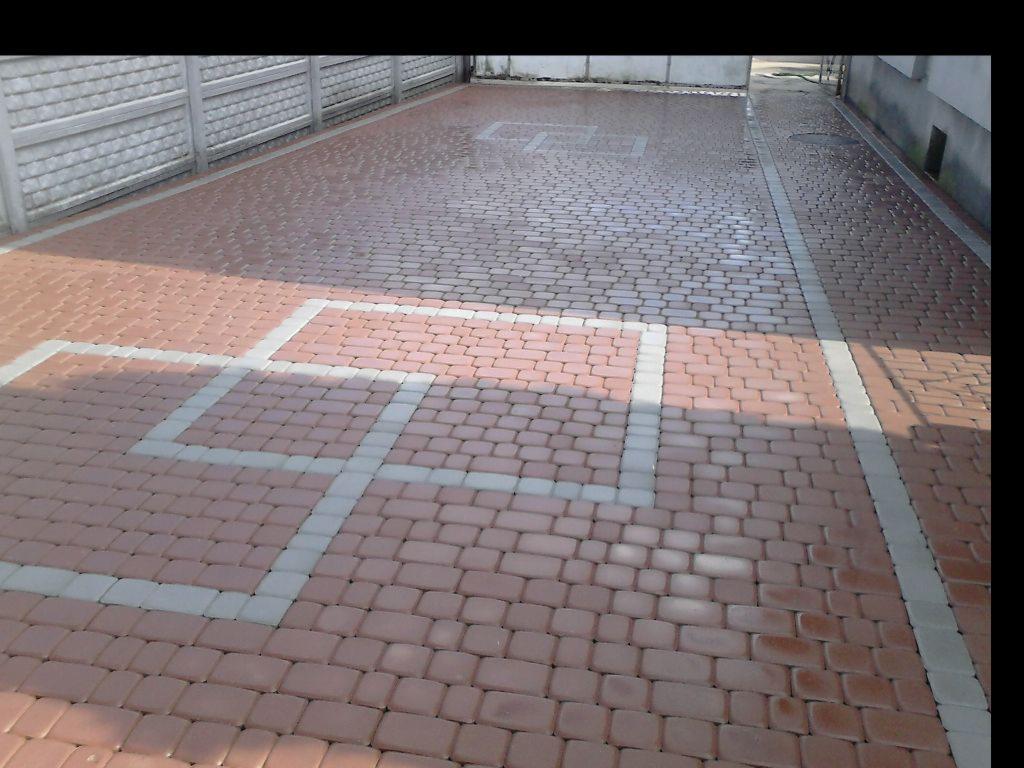 Ułożona kostka brukowa w odcieniach czerwieni i szarości tworzy wzór kwadratów na podjeździe. Szara kostka obramowuje czerwone pola, a całość otacza szary chodnik.