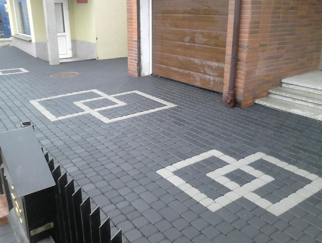 Szara kostka brukowa z geometrycznym wzorem w jasnym odcieniu, ułożona na podjeździe przed budynkiem z brązową bramą garażową i schodami wejściowymi.