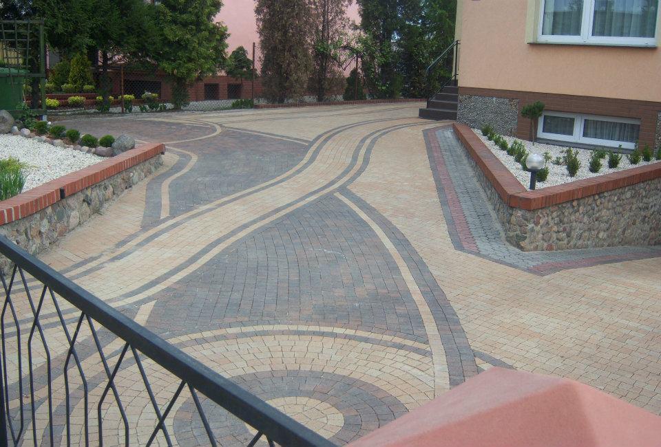 Wzorzysty podjazd z kostki brukowej w odcieniach beżu i szarości, z dekoracyjnymi obrzeżami i niskimi murkami z kamienia i cegły. Widok na fragment domu z oknami.