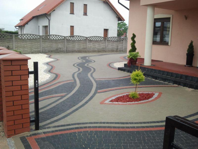 Efektowna nawierzchnia z kostki brukowej ułożona w falujące wzory, z elementami dekoracyjnymi w postaci żwiru i roślinności, prowadząca do domu z kolumną.