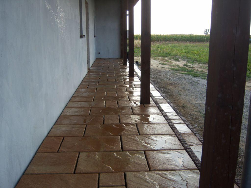 Brązowa kostka brukowa na zewnątrz budynku z drewnianymi słupami. Kostka ułożona w regularny wzór, lekko wilgotna, odbija światło. Tło to trawa i pole.