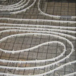 Białe rury ogrzewania podłogowego, ułożone spiralnie na folii z nadrukowaną siatką, zabezpieczone plastikowymi uchwytami.