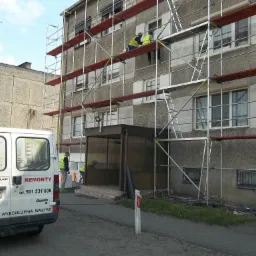 Prace na rusztowaniu przy ocieplaniu elewacji bloku mieszkalnego w Szamotułach, widoczny samochód dostawczy z reklamą usług remontowych.