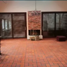 Przestronny salon z terakotową posadzką ułożoną we wzór jodełki, ceglanym kominkiem i minimalistycznym żyrandolem, oświetlony naturalnym światłem wpadającym przez okno i przesuwne drzwi z żaluzjami.