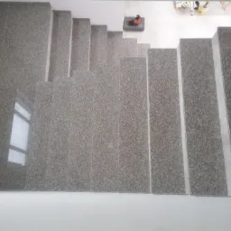 Widok z góry na schody wyłożone szarymi płytkami imitującymi kamień, z widocznym odbiciem okna na pierwszej płytce i narzędziami budowlanymi w tle.