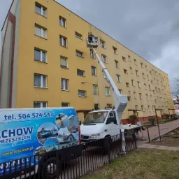 Pracownik w koszu podnośnika czyści elewację bloku mieszkalnego. Na pierwszym planie widoczny samochód dostawczy z reklamą firmy oferującej usługi czyszczenia i malowania.