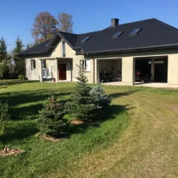 Nowo wybudowany dom jednorodzinny z dwoma garażami i czarnym dachem, widok z trawnika z młodymi drzewkami iglastymi na pierwszym planie.