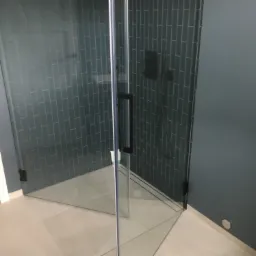 Nowoczesna kabina prysznicowa z grafitową armaturą i płytkami w odcieniach szarości, widok z zewnątrz na fragment łazienki z jasną podłogą.