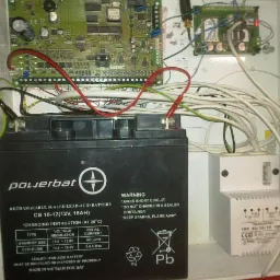 Otwarta skrzynka z elektroniką alarmową: akumulator Powerbat, płytka drukowana i transformator. Widoczne przewody i złącza. Elementy systemu alarmowego.