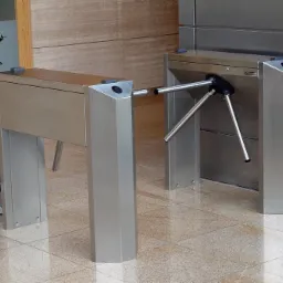 Dwa metalowe, obrotowe systemy kontroli dostępu (tripody) z rośliną w tle, zainstalowane w nowoczesnym biurze. Widok z boku, oświetlenie naturalne.