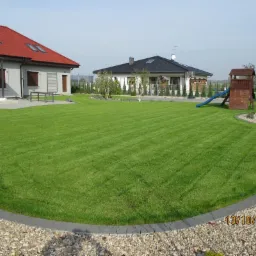 Rozległy trawnik przed domem, z równo skoszoną trawą, obrzeżami z kostki i kamienia. Widoczne dwa budynki mieszkalne i plac zabaw dla dzieci.