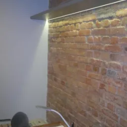 Biuro z ceglaną ścianą, oświetloną półką z segregatorami. Na biurku lampka i kalendarz. Widoczny fragment krzesła. Surowy, loftowy klimat.