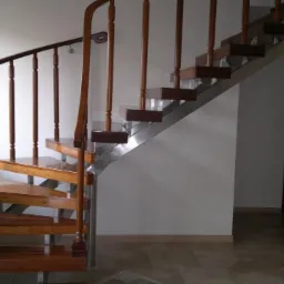 Stylowe, drewniane schody kręte z metalowymi elementami, prowadzące na wyższą kondygnację. Widoczne okno i drzwi w tle. Ujęcie z dołu, ukazujące detale konstrukcji.