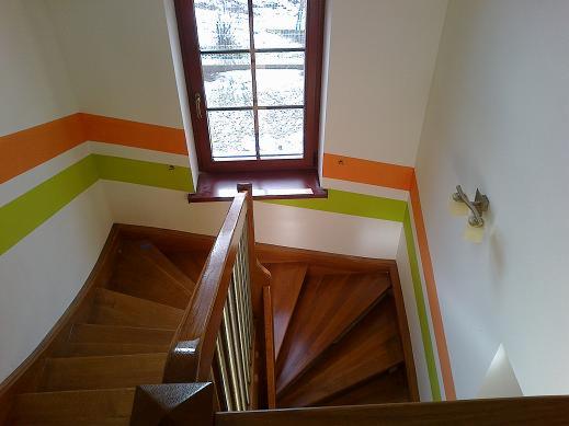 Schody drewniane po remoncie w Będzinie, z pomalowanymi ścianami w pasy zieleni i pomarańczy, widok z góry na klatkę schodową i okno dachowe.