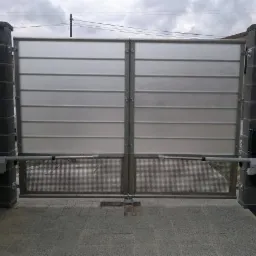 Automatyczna brama wjazdowa z panelem górnym z poziomymi przetłoczeniami i siatkową dolną częścią, zamontowana między kamiennymi słupkami z sygnalizacją świetlną na jednym z nich.