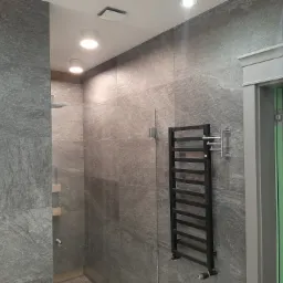 Nowoczesna łazienka wykończona szarymi płytkami imitującymi kamień, z czarnym grzejnikiem drabinkowym i kabiną prysznicową ze szklanymi półkami.