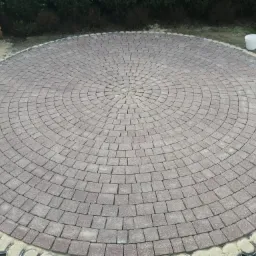 Okrągła powierzchnia wyłożona drobną, szarobrązową kostką brukową ułożoną w koncentryczne wzory, obrzeże z ciemnych elementów, w tle widoczny fragment muru z czerwonej cegły i zieleń.