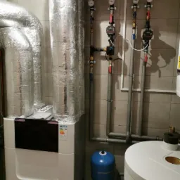 Biały piec gazowy AERIS z czarnym panelem, rury w izolacji aluminiowej, manometry i zawory na szarych rurach, niebieski zbiornik wyrównawczy i biały zasobnik ciepłej wody w kotłowni wyłożonej...