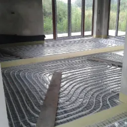 Wnętrze w trakcie instalacji ogrzewania podłogowego: widoczne rury grzewcze ułożone na siatce, zabezpieczone taśmą, surowy beton na ścianach i duże okna z widokiem na zieleń.