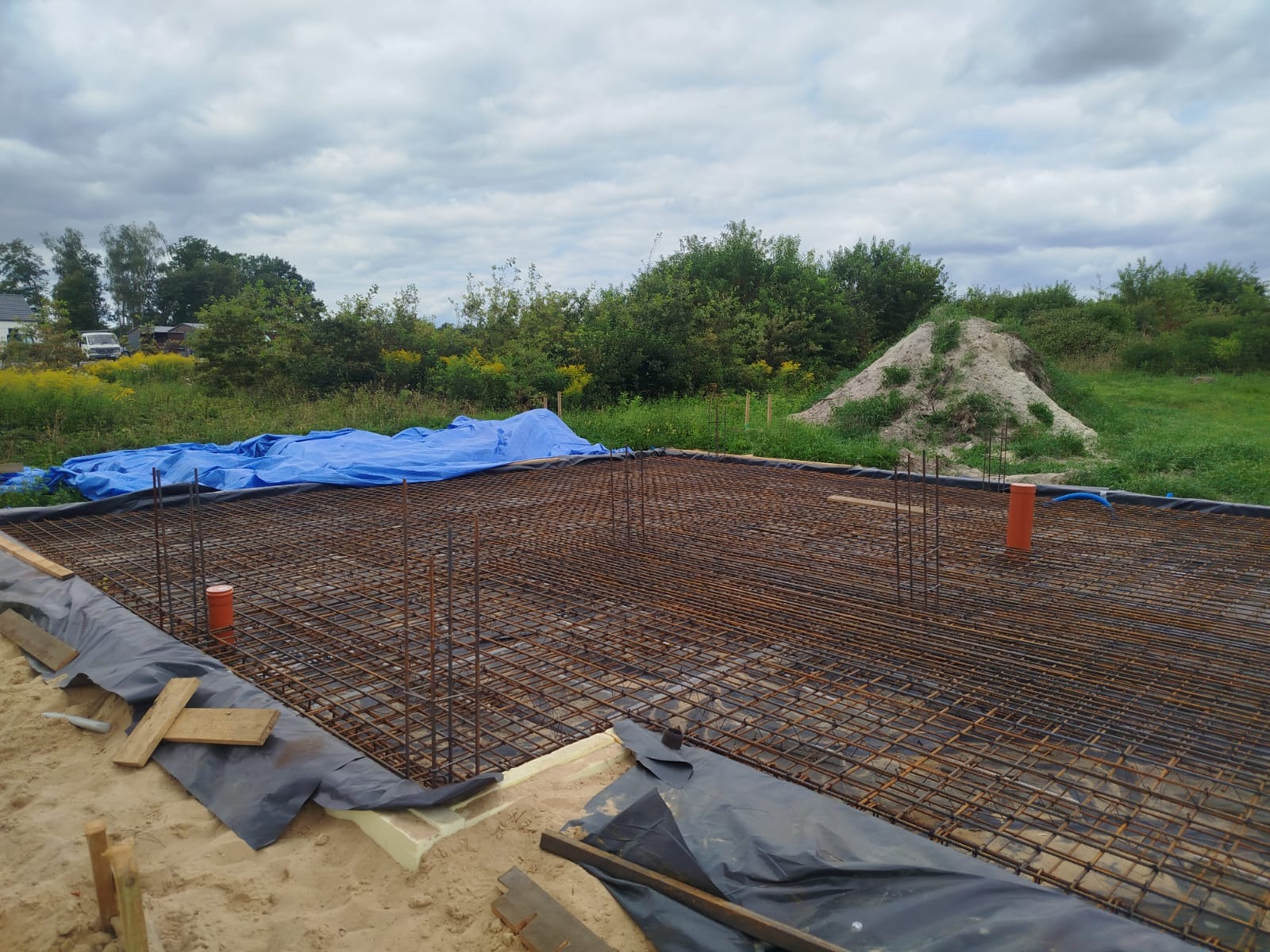Zbrojenie fundamentów domu jednorodzinnego, widoczne pręty zbrojeniowe tworzące siatkę, pionowe słupki, folia izolacyjna i fragment niebieskiej plandeki w tle.